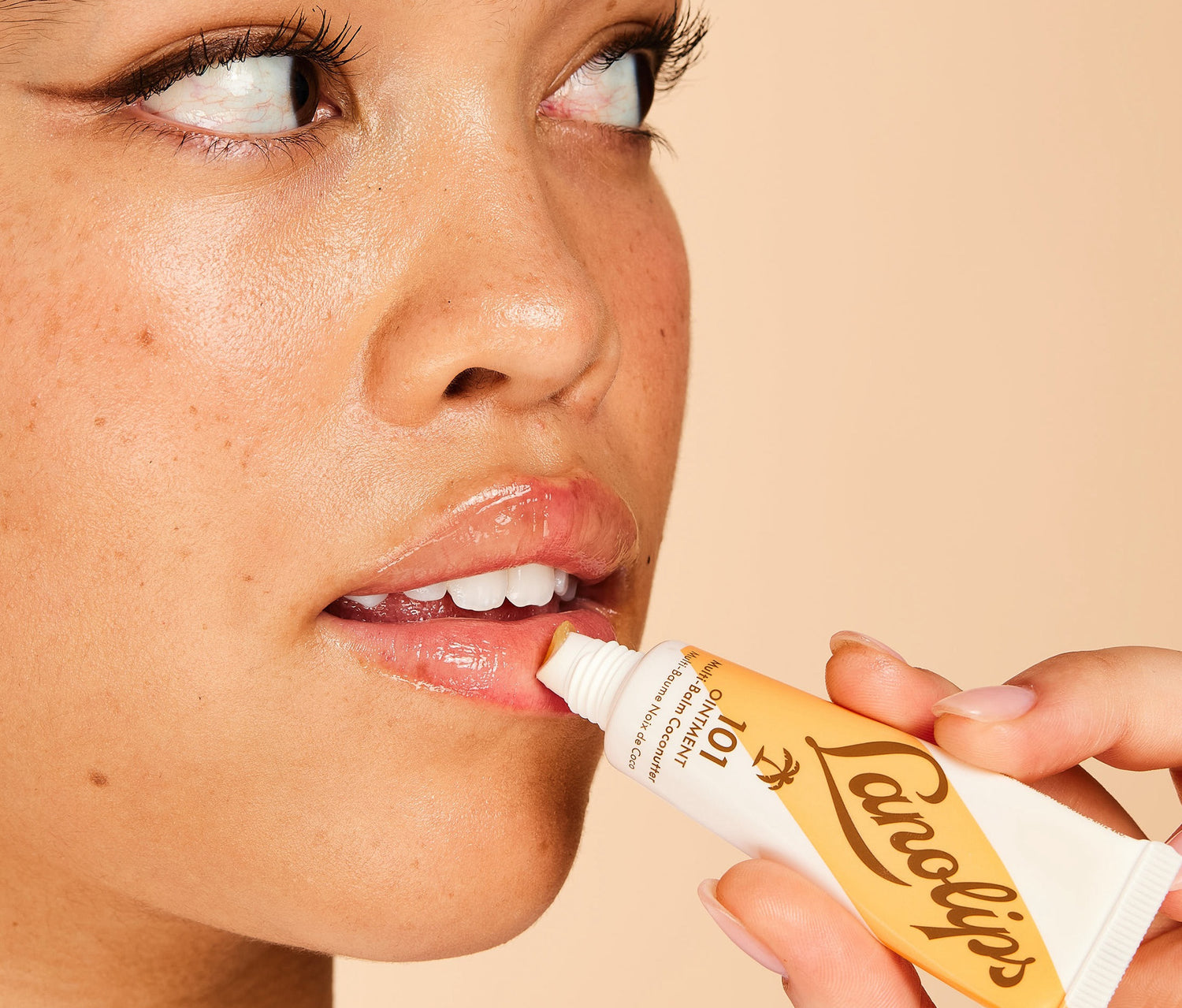 A close up shot of a model applying Lanolips 101 Ointment Multi Balm Coconutter in a caramel coloured tube. Model is pressing the applicator to her lips, looking to the side. The background is a warm peach tone, complementing the golden hues of the tube.  Using ultra-pure grade Aussie lanolin, vitamin e and coconut, his super-rich balm penetrates & seals in moisture to give long-lasting hydration for dry & chapped lips, skin patches, cuticles, elbows & more.