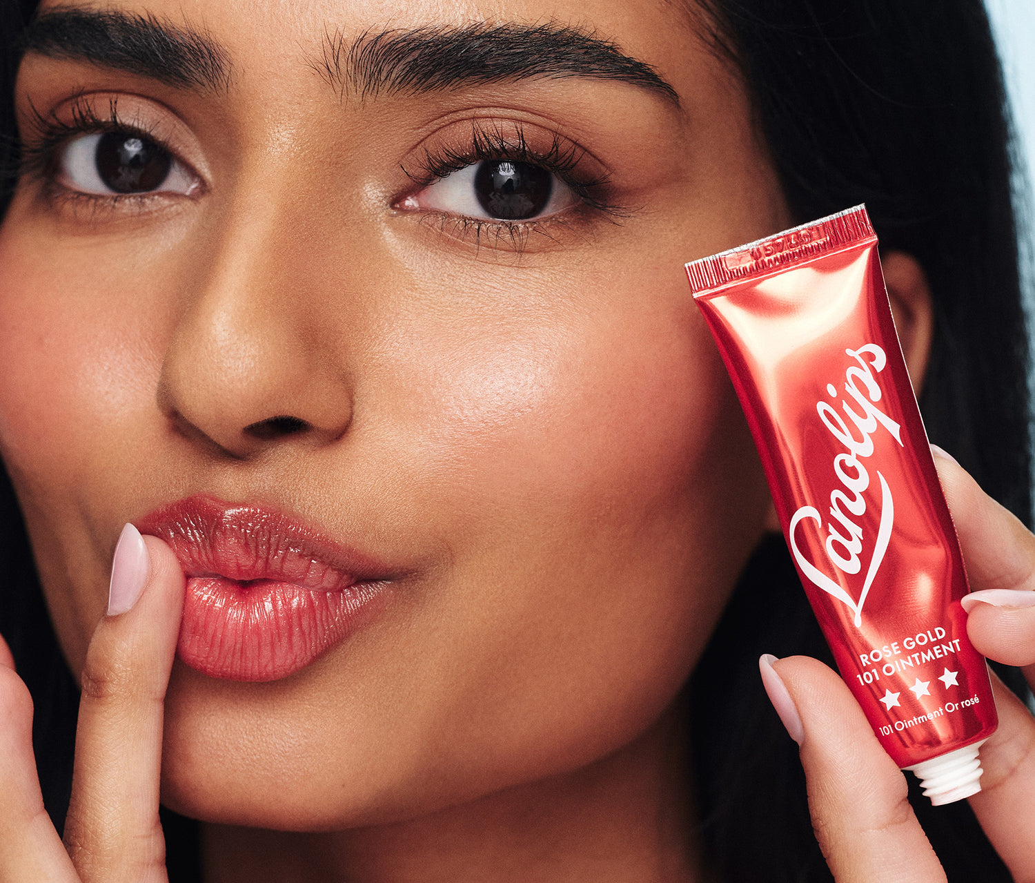 Close-up shot of a model applying Lanolips Rose Gold 101 Ointment to her lips while holding a metallic red tube near her face. This super rich balm, infused with lanolin and a subtle rose gold shimmer, provides deep hydration and a natural rosy flush for both lips and cheeks