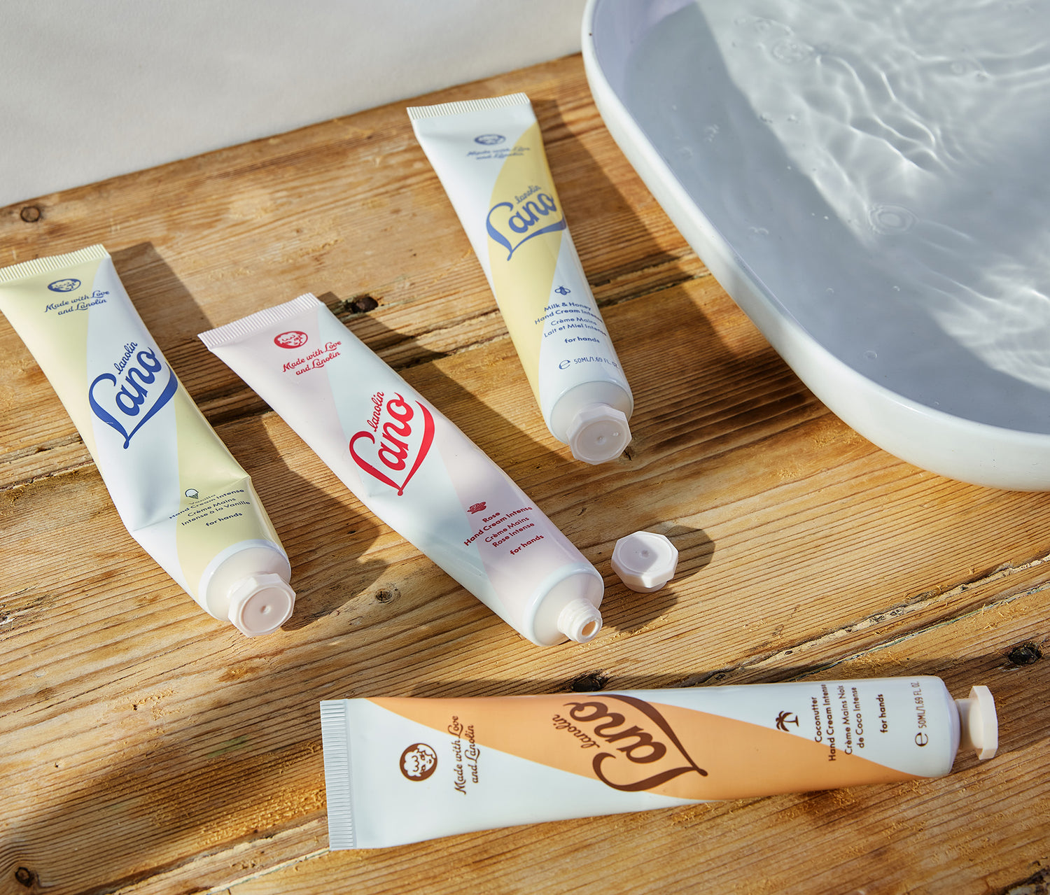 Four tubes of Lanolips Hand Cream Intense rest on a rustic wooden surface beside a bowl of water. Infused with triple lanolin, this rich hand cream deeply nourishes and repairs dry skin, creating a protective moisture barrier. The collection features four indulgent flavours - Coconutter, Rose, Milk & Honey, and Vanilla—each offering long-lasting hydration with a luxurious feel. Perfect for soft, smooth hands, this dermatologically tested formula absorbs beautifully without feeling greasy.