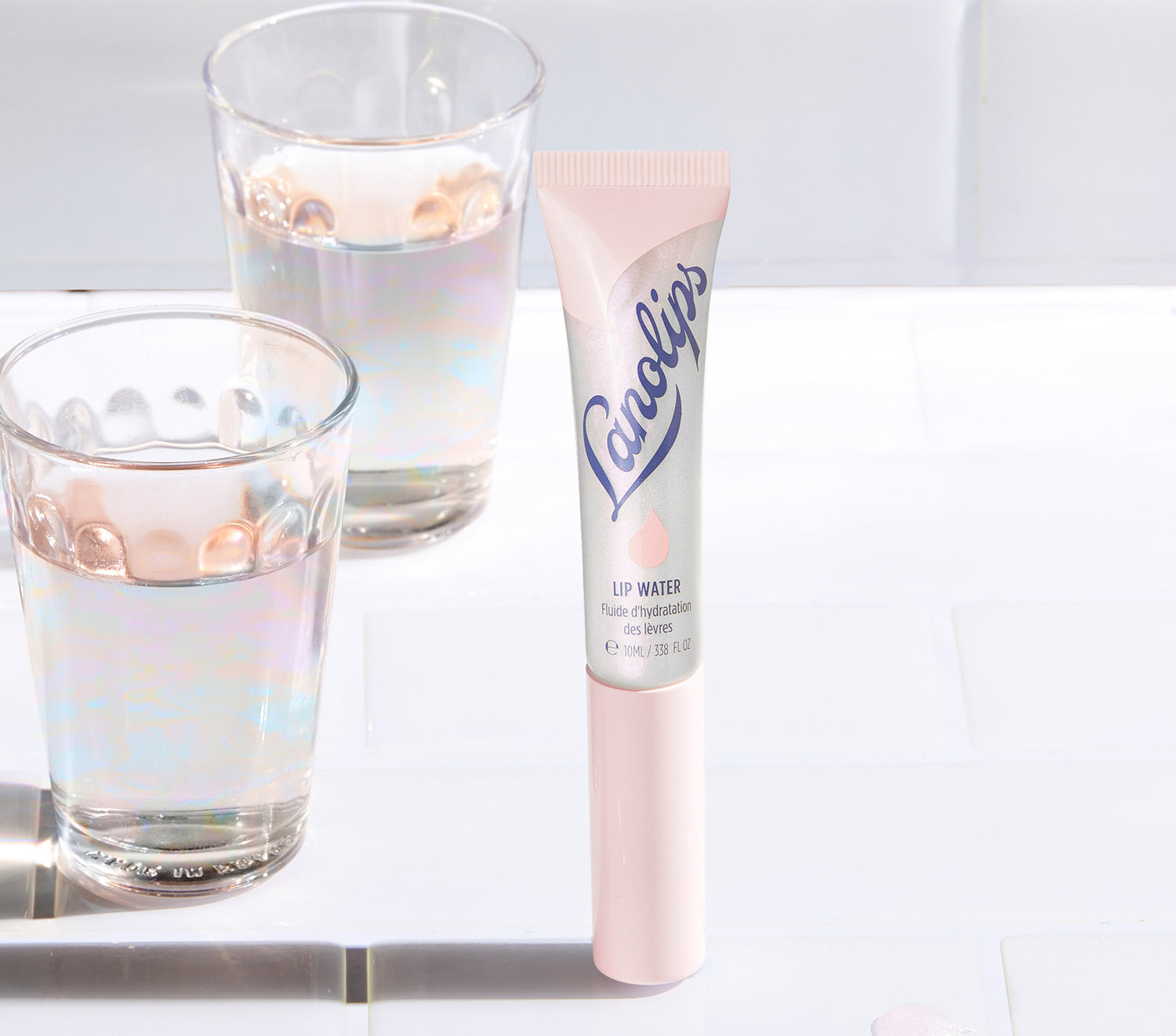 A tube of Lanolips Liquid Lip Water stands on a white surface, with shimmering water glasses in the background. The silver and pink packaging features the blue Lanolips branding and a droplet icon. Made with liquid lanolin, hyaluronic acid, glycerin and peppermint, this non-sticky, hydrating lip serum delivers deep moisture with a underwater holographic glow. Like a drink of water for your lips, it nourishes, softens and adds a glass like sheen - perfect for dry lips and lip lickers.