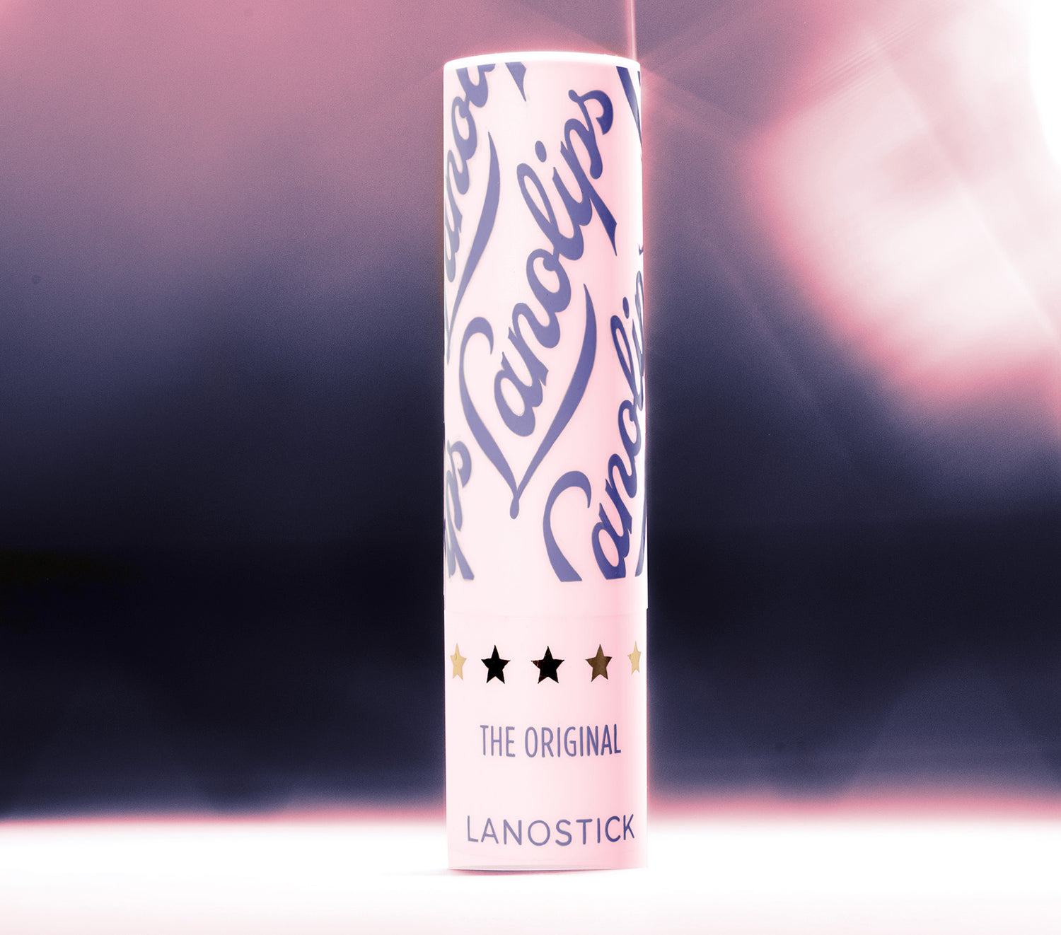 A sleek tube of Lanolips Lanostick stands against a soft pink and black gradient background. This 90s-inspired wind-up lip balm is perfect for pockets, bags, and on-the-go hydration. From the makers of the 'World’s Best Lip Balm,' Lanostick glides on smoothly, delivering a rich layer of moisture that deeply conditions lips. Infused with lanolin, natural waxes, butters, and vitamin E, it nourishes and locks in hydration for hours. The ultimate day-balm for soft, healthy lips.