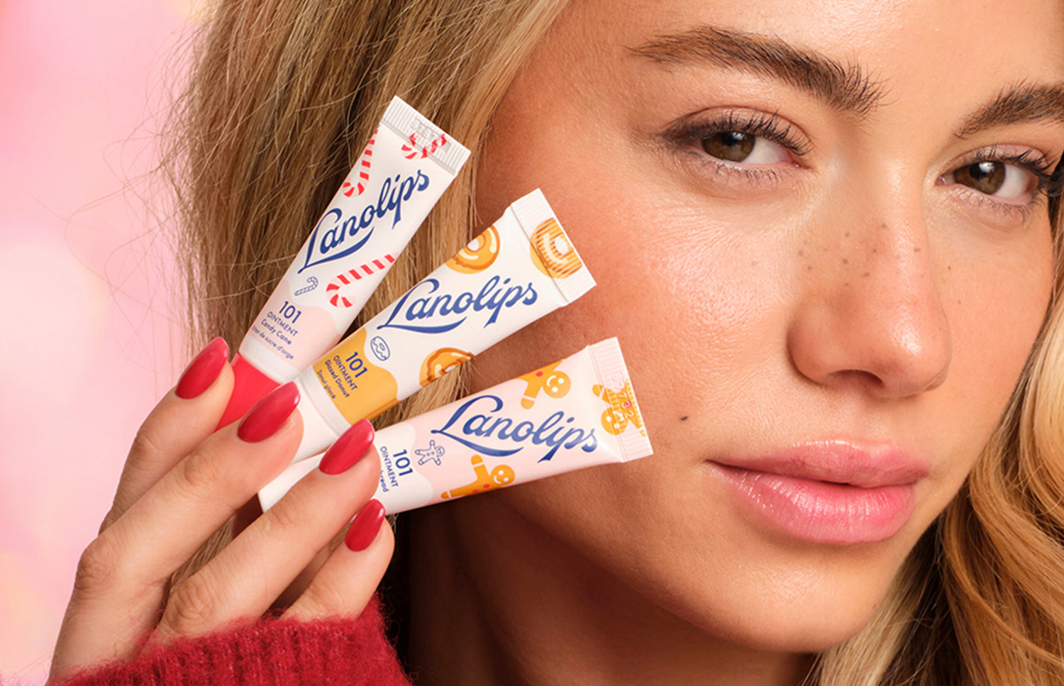 Model holding mini Lanolips lip balm tubes Candy Cane, Glazed Donut and Gingerbread in front of her face against a pink background. The minis are part of the Lanolips Holiday Mini Trio.