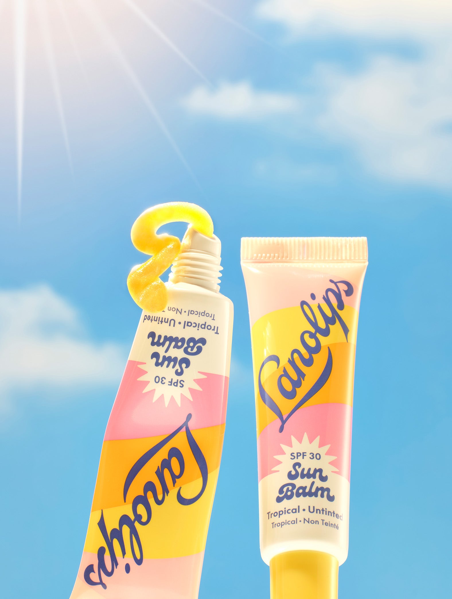 Set against a radiant blue sky with a shining sun, two tubes of Lanolips Sun Balm SPF 30 Tropical lip balm are featured - one standing upright and the other squeezed to reveal a vibrant, glossy balm shaped like a “3” to highlight its SPF 30 protection. This creamy, radiant formula is packed with hydrating lanolin, SPF
protection, and a luminous sheen.