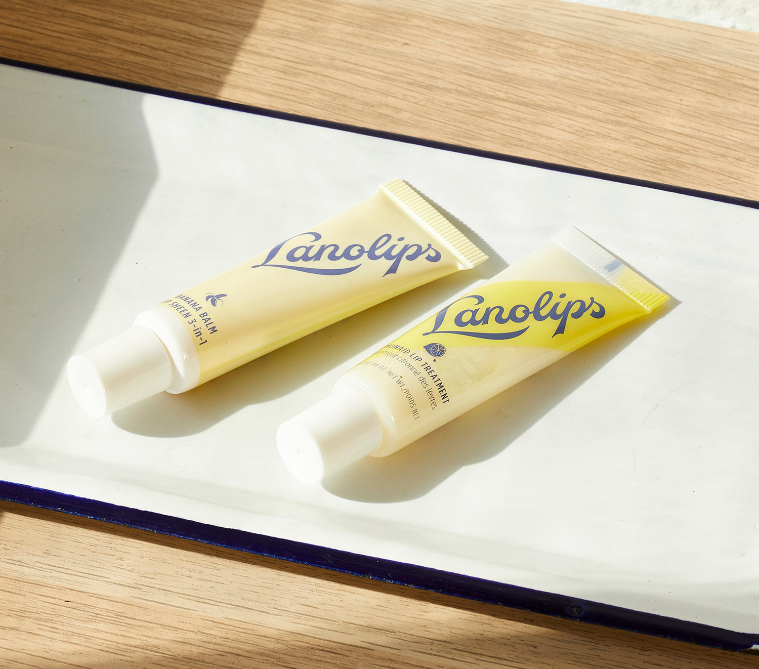 Two tubes of Lanolips Banana Balm Lip Sheen and Lemonaid Lip Treatment are displayed on a ceramic tray in natural sunlight. This best-selling lip duo includes two full-size treatments made with ultra-pure lanolin. Banana Balm is a creamy 3-in-1 formula that acts as a lip conditioner, gloss, and protectant, with added banana extract for soft, glowing lips. Lemonaid Lip Treatment features whipped lanolin and all-natural lemon oil to exfoliate and hydrate, leaving lips smooth, fresh, and deeply moisturised.