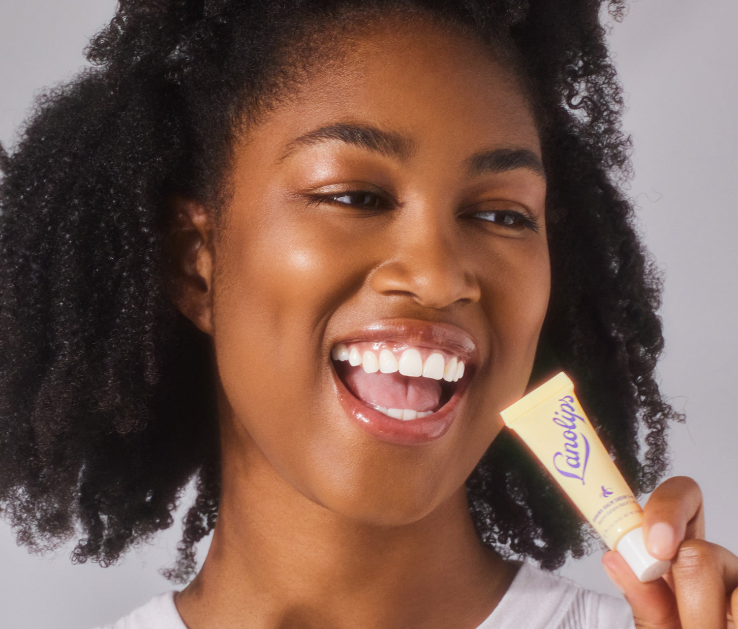 A model holding a tube of Lanolips Banana Balm Sheen 3-in-1 ner her face. Her lips are coated with the lip sheen, providing deep hydration and a subtle sheen. Made with lanolin, banana extract, cocoa seed butter and a hint of shimmer, this balm nourishes while delivering a soft, fruity scent. 