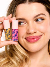 Load image into Gallery viewer, Smiling model holding two Lanolips Hyaluronic Lip Oil shades, highlighting the product&#39;s sleek pink packaging. The lip oils, infused with lanolin, hyaluronic acid, and vitamin E, deliver a glossy and nourishing finish for soft, hydrated lips
