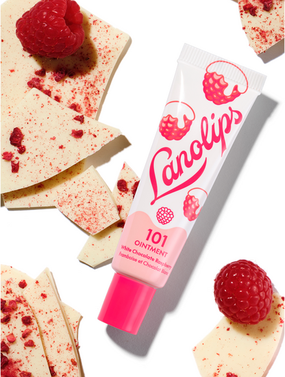 Lanolips 101 Ointment Multi-Balm White Chocolate Raspberry 101 Ointment from the 101 Delicious collection. A close-up shot of the multi-balm tube nestled among creamy white chocolate shards dusted with dried raspberry and fresh, whole raspberries.
