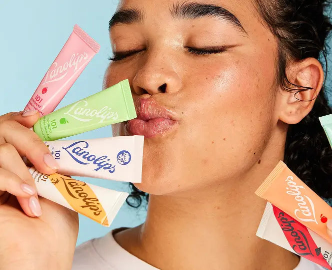 Close-up of a model holding several products from the Lanolips 101 Ointment range, including The Original 101 Oitnment Multipurpose Superbalm, Coconutter, Strawberry, Green Apple, Peach, and Watermelon. These cult-favourite, multi-award-winning balms have been voted ‘The World’s Best’. Powered by ultra-pure lanolin, they intensely hydrate and nourish dry lips and skin. Hydration is the foundation of skincare  - start with lanolin.