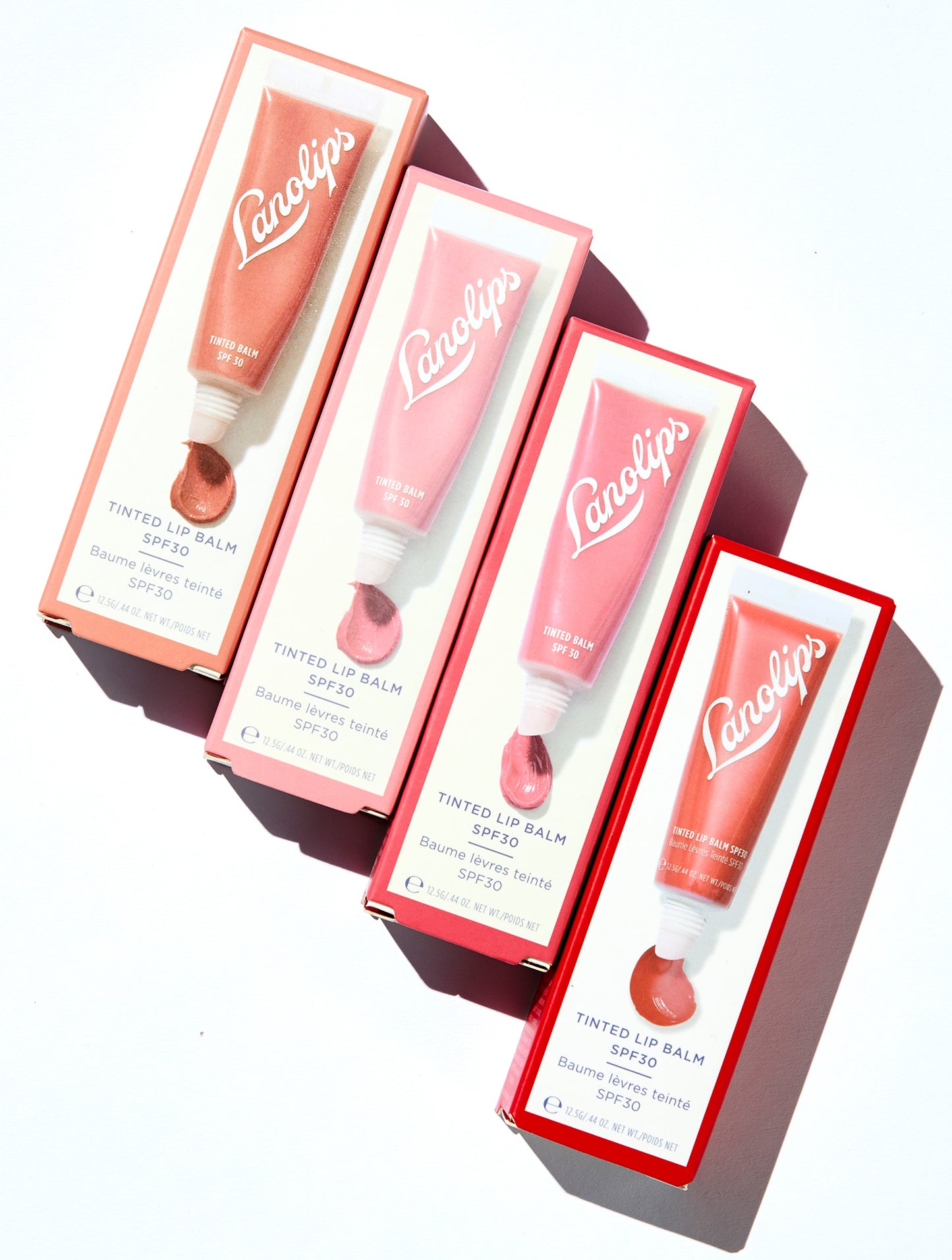 Flat lay of the Lanolips Tinted Lip Balm boxes in four shades - Rose, Rhubarb, Perfect Nude, and Red Apple - arranged in a geometric stack casting a soft shadow. This hydrating balm adds a natural tint while nourishing lips with lanolin. Ideal for everyday use and dry lip care.