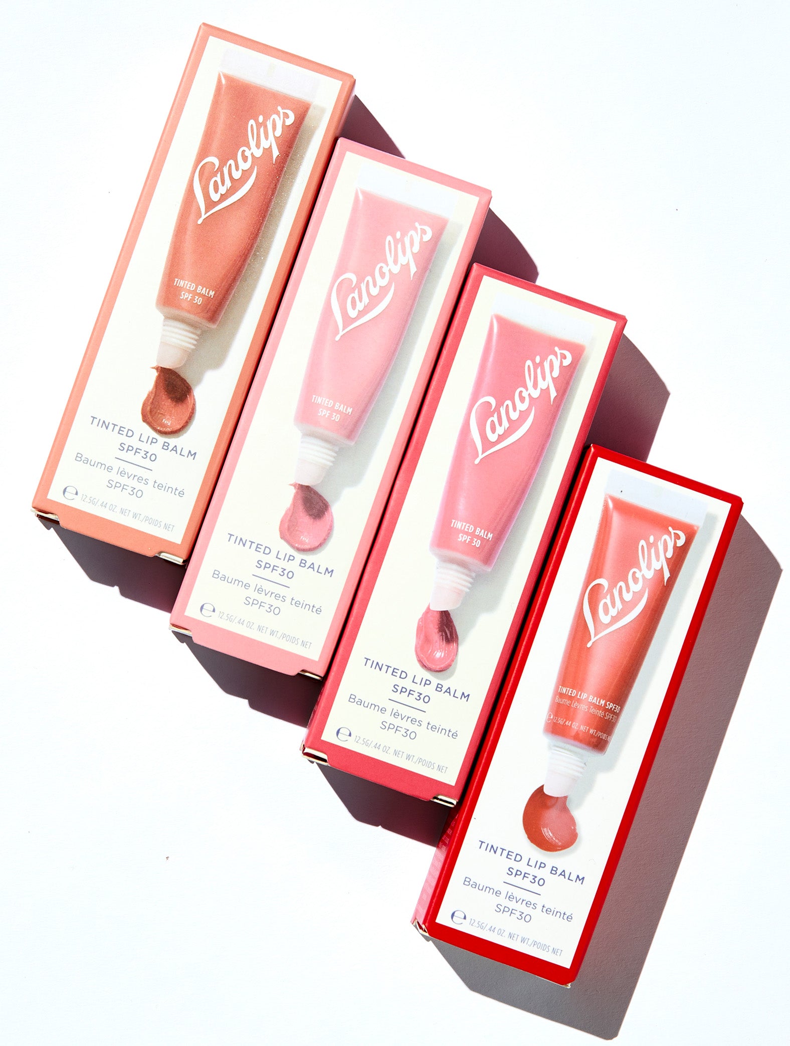 Flat lay of the Lanolips Tinted Lip Balm boxes in four shades - Rose, Rhubarb, Perfect Nude, and Red Apple - arranged in a geometric stack casting a soft shadow. This hydrating balm adds a natural tint while nourishing lips with lanolin. Ideal for everyday use and dry lip care.