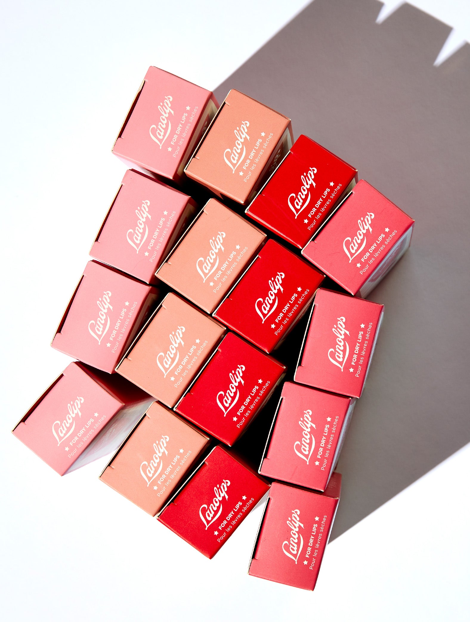Flat lay of Lanolips Tinted Lip Balm SPF30 boxes in four shades - Rose, Rhubarb, Perfect Nude, and Red Apple - arranged in a geometric stack casting a soft shadow. This hydrating balm adds a natural tint while nourishing lips with lanolin. Ideal for everyday use and dry lip care.