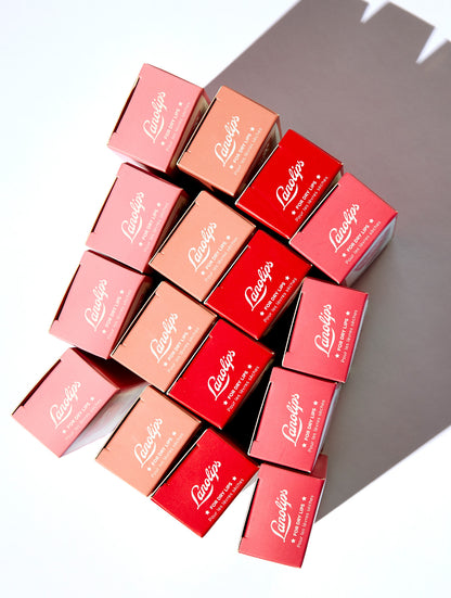 Flat lay of Lanolips Tinted Lip Balm SPF30 boxes in four shades - Rose, Rhubarb, Perfect Nude, and Red Apple - arranged in a geometric stack casting a soft shadow. This hydrating balm adds a natural tint while nourishing lips with lanolin. Ideal for everyday use and dry lip care.