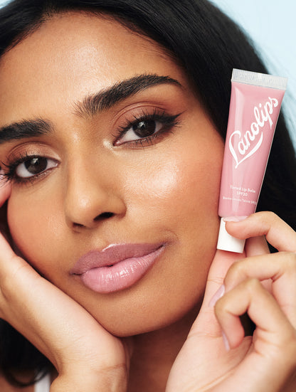 Close up of model wearing the Lanolips Tinted Lip Balm SPF30 Rose on her lips. She also holds the tube of the Tinted Lip Balm in Rose, a creamy tea pink shade designed to enhance natural lip colour. This cult-favourite formula is made with lanolin, vitamin e, shea butter and SPF30 for a long-lasting, non-sticky finish. With the right amount of pigment and high-gloss finish, it's the perfect everyday lip balm. The SPF30 helps shield lips from sun damage. 