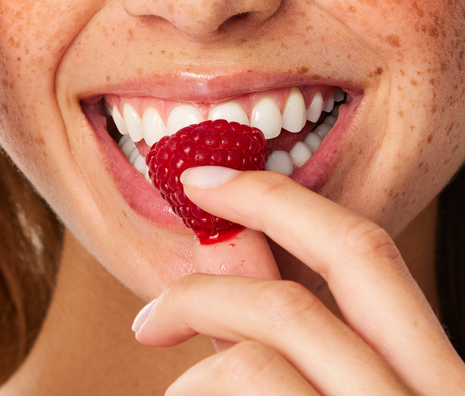 A macro close-up of a fresh, juicy raspberry being held against smiling lips to highlight the natural inspiration behind Lanolips 101 Ointment Multi-Balm in White Chocolate Raspberry. Part of the 101 Delicious collection.